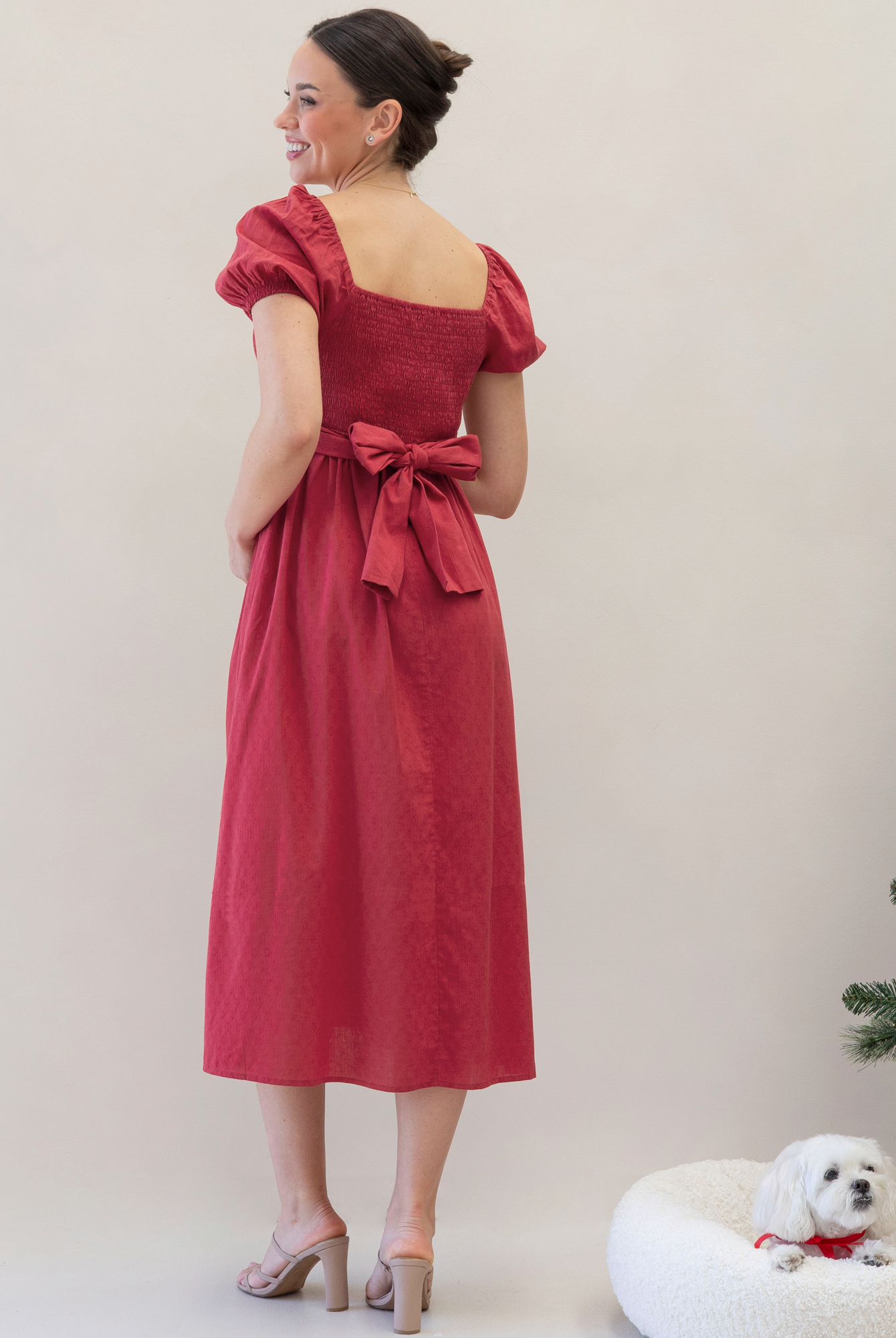 Woman in a red dress with a bow standing next to a small white dog on a white background