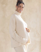 A pregnant woman wearing a cream-colored maternity turtle neck knitted sweater with oversized sleeves.