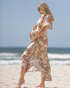 A pregnant woman standing on a beach wearing a beige maternity dress with a palm leaf print, a V-neck, and flutter sleeves.