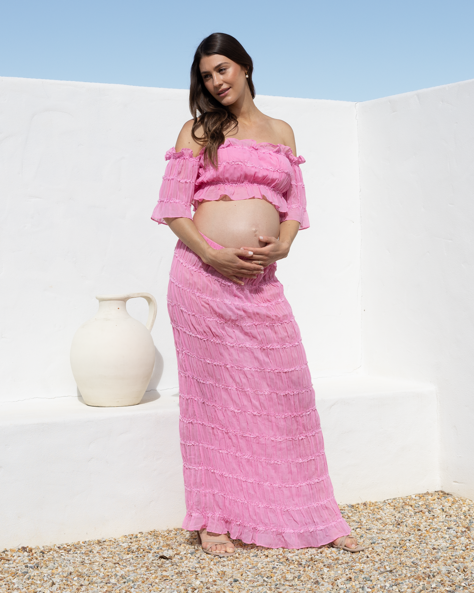 A pregnant woman wearing a pink off-the-shoulder two-piece set consisting of a cropped top and a long maxi skirt, standing outdoors against a white stucco wall.