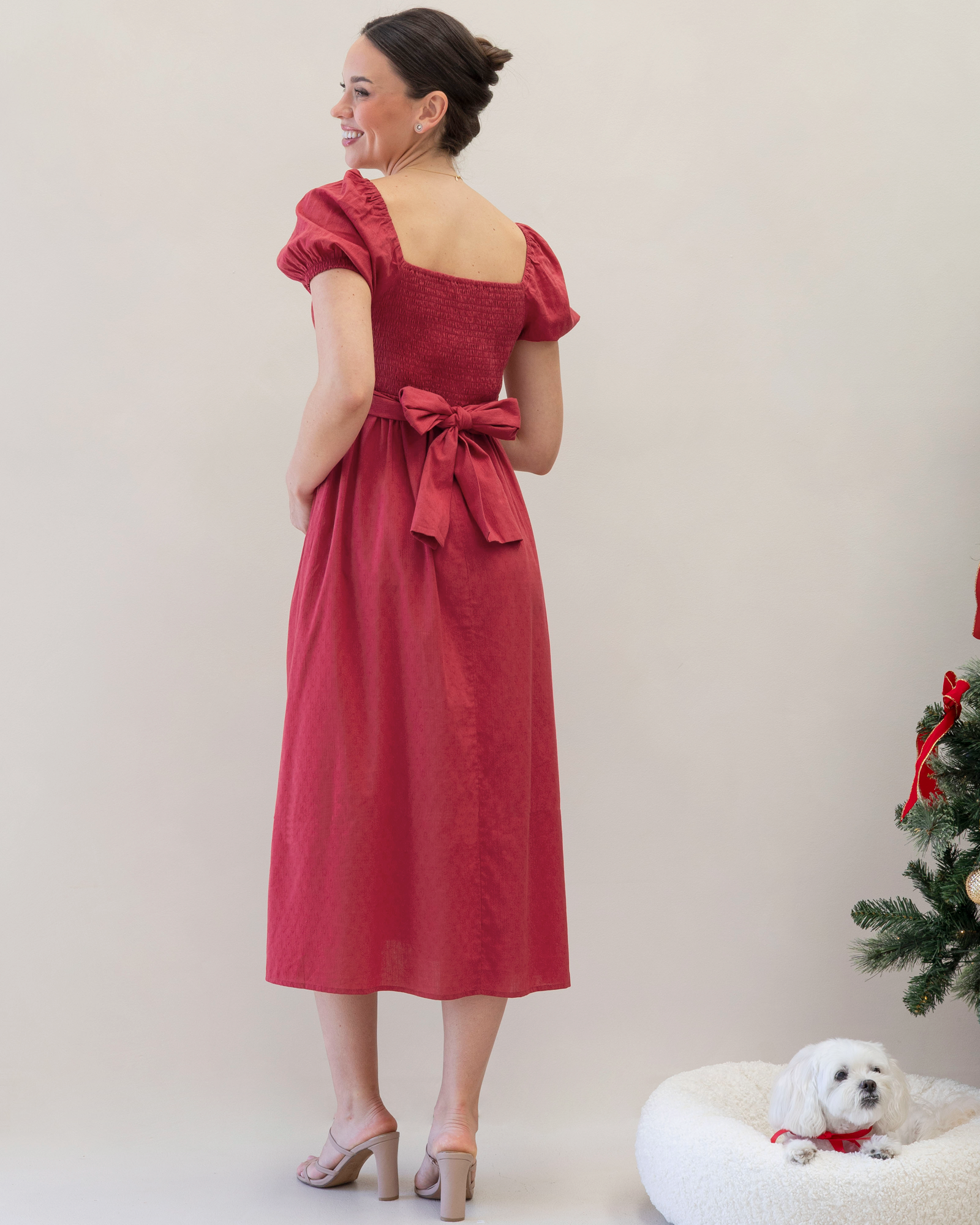 Woman in a red dress with a bow standing next to a small white dog on a white background
