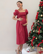 Woman in a red dress standing next to a decorated Christmas tree with a small dog on a white cushion.