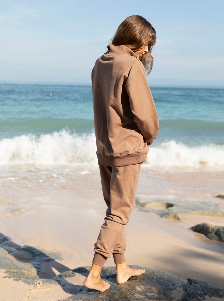 Back view - A pregnant woman in 2-Piece Calla zip front brown Maternity Tracksuit Set walking on a beach (6648836292702)