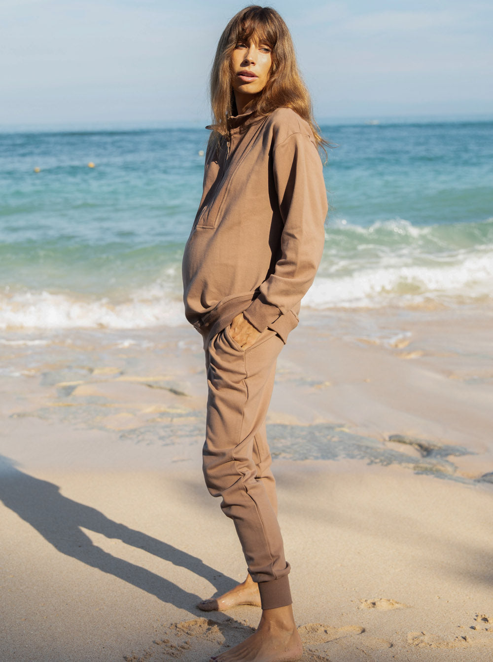 Side view - A pregnant woman in 2-Piece Calla zip front brown Maternity Tracksuit Set standing on a beach (6648836292702)