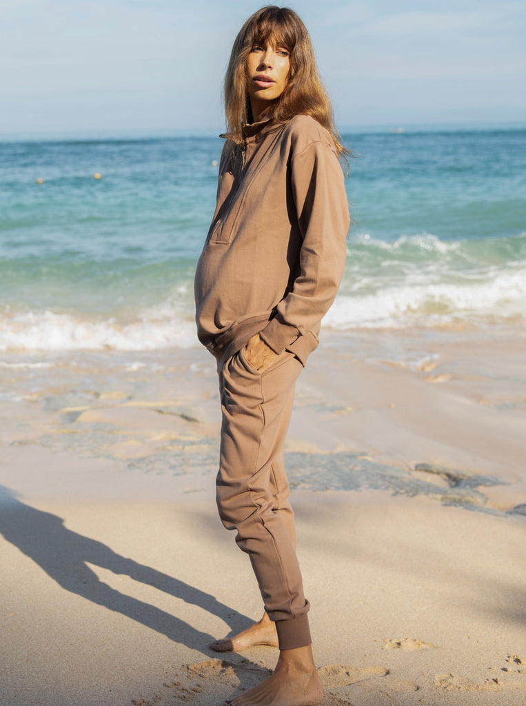 Side view - A pregnant woman in 2-Piece Calla zip front brown Maternity Tracksuit Set standing on a beach (6648836292702)